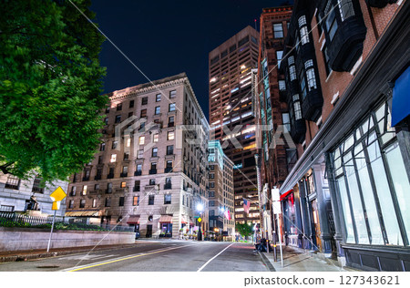 Night scene in Boston, Massachusetts, United States, shows a quiet downtown street lined by historic brick buildings and a modern high rise, tree foliage glowing under streetlights and dark sky. Night scene in Boston, Massachusetts, United States, shows a quiet downtown street lined by historic brick buildings and a modern high rise, tree foliage glowing under streetlights and dark sky. 127343621