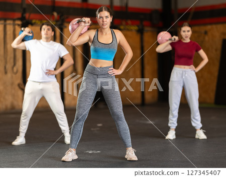 Girl in gym does exercises with kettlebell, lifts and lowers heavy kettlebell with one hand 127345407