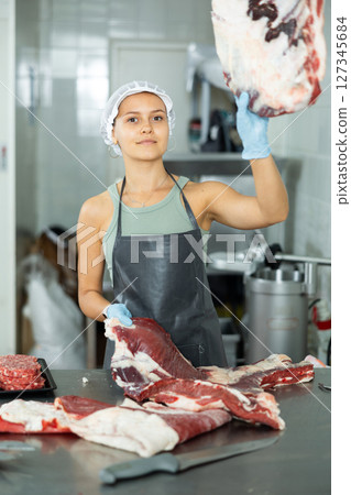 Girl in butcher shop cuts meat carcass into small pieces of meat for retail sale in small store 127345684