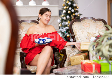 Smiling young female in red dress examining packed gift box with bow on sofa in front of Christmas tree at apartment 127345688