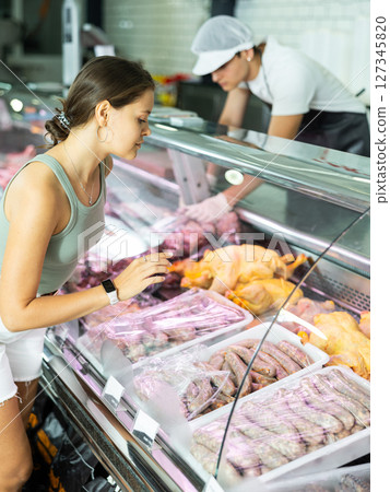 Girl buyer of meat department store, looking at meat small sausage in refrigerator window 127345820