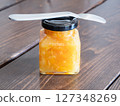 A glass jar and spoon filled with marmalade jam, made by sweetly stewing citrus peels and fruits. 127348269