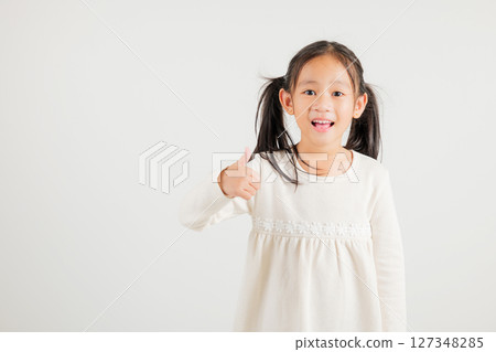 Confident Asian young girl in a studio portrait isolated on white background makes a thumbs up OK sign, symbolizing agreement and success. kindergarten child smile radiates positivity and satisfaction 127348285