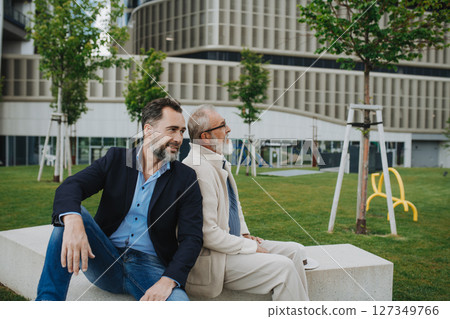 Father and adult son sitting on bench in park, sharing peaceful moment. 127349766