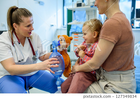 Pediatrician demonstrates oxygen mask on teddy bear to young patient 127349834
