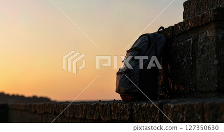 Silhouetted Backpack at Sunrise on a Stone Wall in Scenic Travel Destination Silhouetted Backpack at Sunrise on a Stone Wall in Scenic Travel Destination 127350630