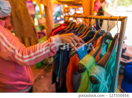 Local Thai market visitor browsing variety of colorful traditional clothing and garments displayed by rural vendors offering authentic handcrafted apparel for tourists and cultural shoppers 127350956