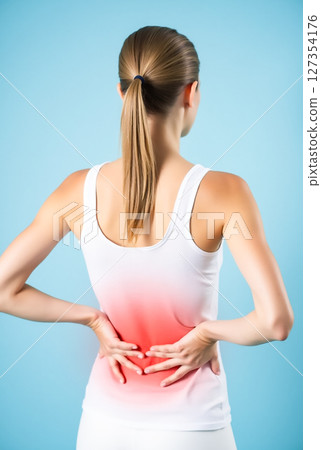 Woman experiencing lower back pain while standing against a light blue background during daylight hours 127354176