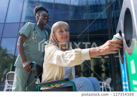 Woman on a wheelchair throwing waste to the bin, nurse carrying her wheelchair 127354389