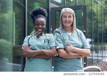 Smiling female doctors in lab coats standing near clinic and looking confident Smiling female doctors in lab coats standing near clinic and looking confident 127354395