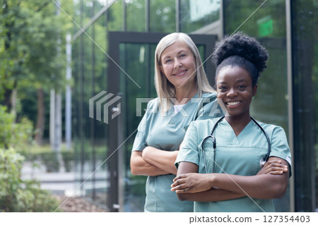 Smiling female doctors in lab coats standing near clinic and looking confident Smiling female doctors in lab coats standing near clinic and looking confident 127354403