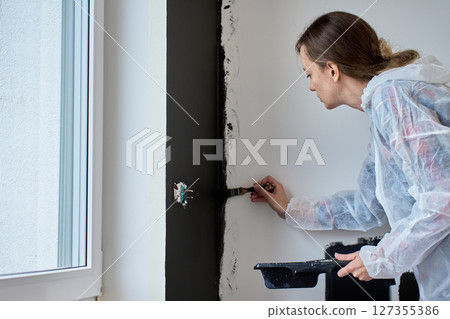 Woman in protective suit painting wall black during apartment renovation using brush Woman in protective suit painting wall black during apartment renovation using brush 127355386