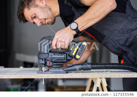Man Cutting Wood Panel with Electric Jigsaw in Workshops 127355546