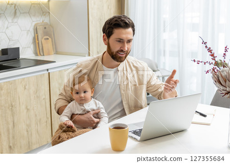 Bearded young man sitting at the laptop and holding his baby boy Bearded young man sitting at the laptop and holding his baby boy 127355684