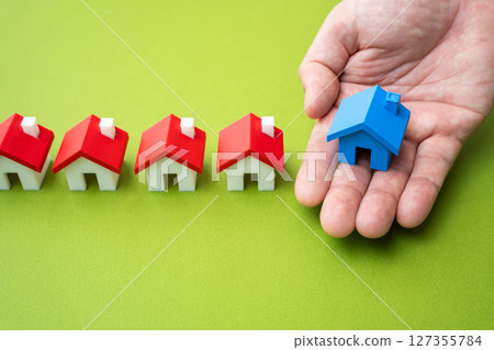 The hand delivers the blue house to the row of others. Buy or sell a home. Property purchase, sale, or selection in the housing market. The hand delivers the blue house to the row of others. Buy or sell a home. Property purchase, sale, or selection in the housing market. 127355784
