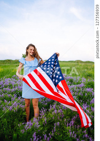 Young woman proudly hold waving american USA flag on blooming meadow. 4th of July. Independence Day. Young woman proudly hold waving american USA flag on blooming meadow. 4th of July. Independence Day. 127356939