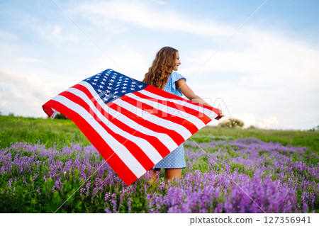 Young woman proudly hold waving american USA flag on blooming meadow. 4th of July. Independence Day. 127356941