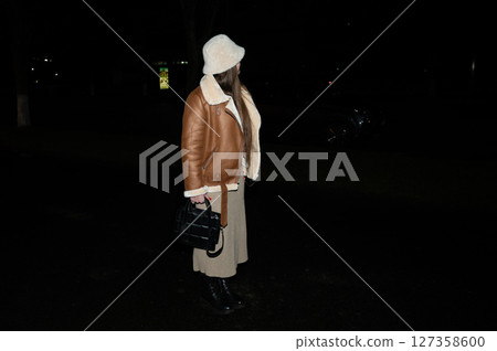 Stylish night walk featuring cozy brown jacket and long skirt in a city park, illuminated by distant streetlights on a crisp evening Stylish night walk featuring cozy brown jacket and long skirt in a city park, illuminated by distant streetlights on a crisp evening 127358600