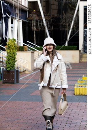 Charming woman in cozy sweater dress and stylish jacket strolls through modern urban setting on a crisp sunny day 127358608