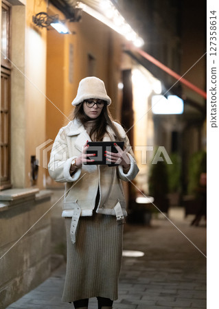 Elegant evening stroll through cobbled streets with a fashionable woman holding a tablet in warm winter attire amidst charming city lights 127358614