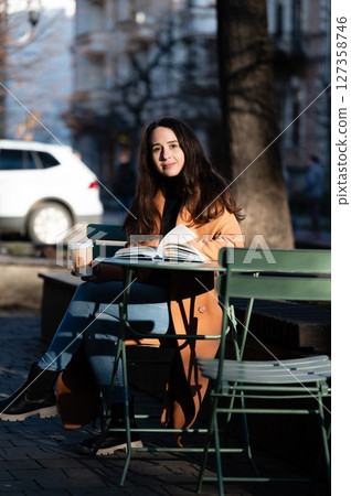 Sunlit afternoon at a city cafe with a young woman enjoying a book and coffee in a peaceful outdoor setting surrounded by urban charm 127358746