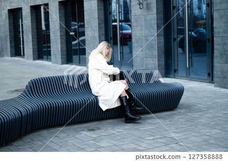 Chic woman sitting on a modern wave-shaped bench while adjusting her stylish bag in a sleek urban environment during chilly afternoon hours 127358888