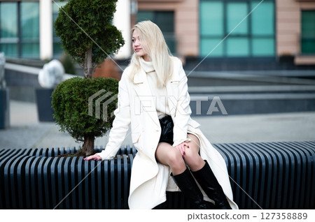 Charming woman in white coat enjoys a stylish moment in an urban park setting, contrasting natural greenery with modern architecture during a crisp afternoon 127358889