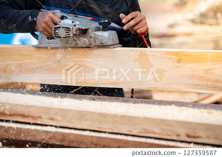 Male carpenter hand using an electric planer on wooden plank at construction site 127359587