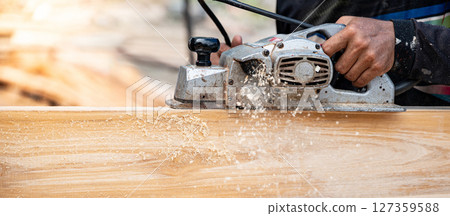 Male carpenter hand using an electric planer on wooden plank at construction site 127359588