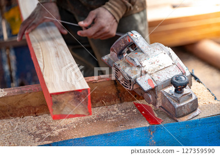 Rustic electric planer on workbench with carpenter hand squaring timber behind 127359590
