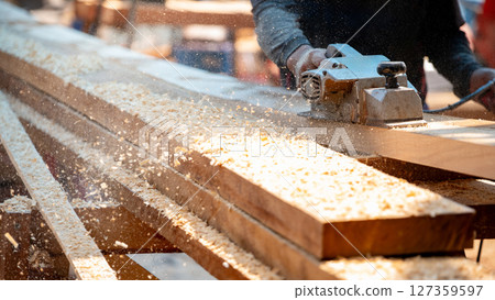 Male carpenter hand using an electric planer on wooden plank at construction site 127359597