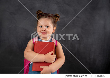 Cute little school girl with book smiling on black chalkboard. 127360029