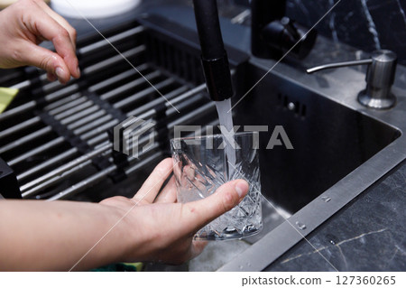 A Person Washing a Textured Glass in a Modern Kitchen Sink Under Running Water Captured in an Elegant and Clean Atmosphere for Optimal Hygiene and Home Maintenance 127360265