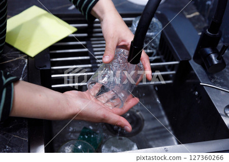 A Clear Glass Being Washed Under a Faucet in a Modern Kitchen Sink Capturing the Careful Attention to Dishwashing with Water Splashes Highlighted and Surrounding Kitchen Details 127360266