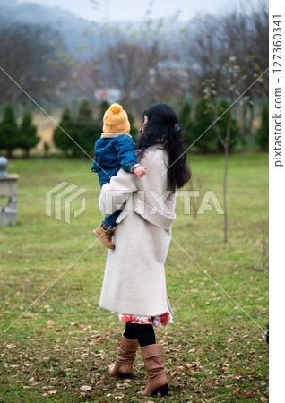 Gentle embrace in autumnal air mother carries her toddler son through tranquil grounds on a crisp afternoon of shared warmth and love as leaves gently rustle Gentle embrace in autumnal air mother carries her toddler son through tranquil grounds on a crisp afternoon of shared warmth and love as leaves gently rustle 127360341