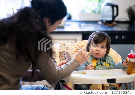 Early nourishment an adorable young child eats pumpkin puree from a spoon at the kitchen table while his mother feeds him and a cup is nearby Early nourishment an adorable young child eats pumpkin puree from a spoon at the kitchen table while his mother feeds him and a cup is nearby 127360343