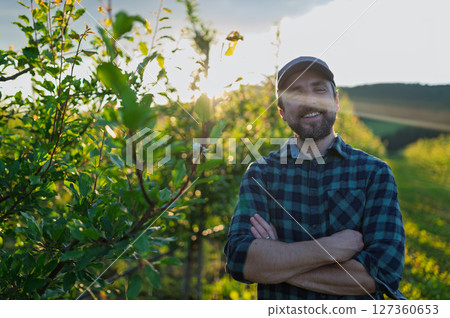 A mature farmer standing in the middle of orchard at sunset. Copy space. A mature farmer standing in the middle of orchard at sunset. Copy space. 127360653