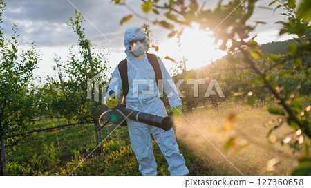 Farmer spraying pesticide in orchard, disinfecting trees in protective suit. 127360658