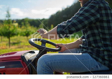 Close up of farmer driving small tractor in orchard, enjoying peaceful moment. Close up of farmer driving small tractor in orchard, enjoying peaceful moment. 127360861