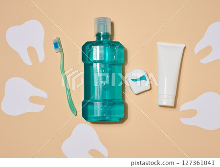 Oral hygiene set, mouthwash, toothbrush, toothpaste and dental floss, surrounded by paper cutout teeth silhouettes on beige background. 127361041