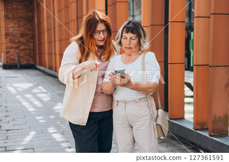 Smiling happy cool fun elder parent mom with young adult daughter two women together wear casual clothes hold in hand use mobile cell phone isolated on urban background. Family day concept Smiling happy cool fun elder parent mom with young adult daughter two women together wear casual clothes hold in hand use mobile cell phone isolated on urban background. Family day concept 127361591