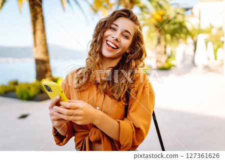 Portrait of a young woman holding a phone in her hands on a city avenue, on a sunny day. 127361626