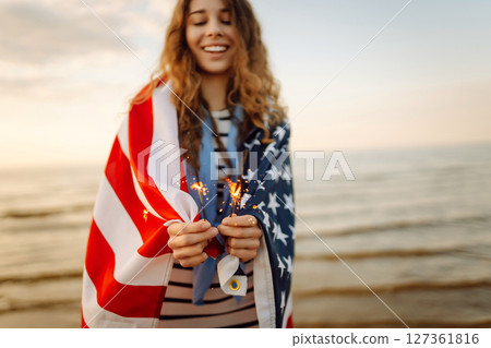 Young woman with American flag and sparklers on beach. Patriotic holiday. USA celebrate 4th of July. 127361816