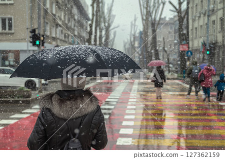 Woman with black umbrella and fur coat waiting on crosswalk during snowfall in city. Dark silhouette contrasts with snowy urban background. Concept of solitude, cold weather routine, city life 127362159