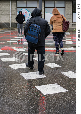 People in winter jackets crossing wet crosswalk during snowfall, urban cold weather. City street with puddles and motion, winter routine. Concept of everyday commute, winter street life, casual day 127362161