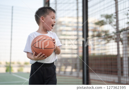 Young boy joyfully playing basketball on a sunny outdoor court. 127362395