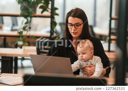 Sitting by laptop. Young mother with her little baby son is in the office 127362531