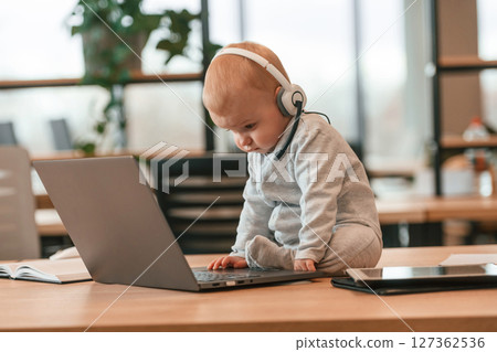 In headphones. Cute little baby boy is in the modern office on the table by laptop In headphones. Cute little baby boy is in the modern office on the table by laptop 127362536