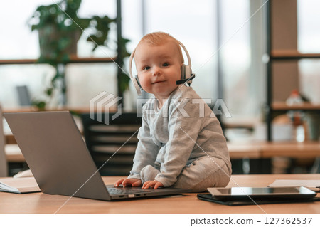 In headphones. Cute little baby boy is in the modern office on the table by laptop In headphones. Cute little baby boy is in the modern office on the table by laptop 127362537