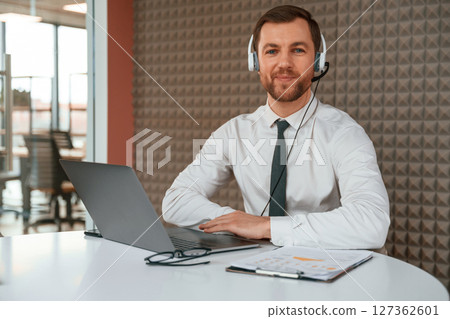 In the headphones. Handsome man in formal clothes is working by laptop in the office 127362601